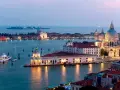 La **Punta della Dogana** a Venezia, con la Basilica di Santa Maria della Salute sullo sfondo, illuminata al crepuscolo. Barche e yacht ormeggiati nell'acqua calma.