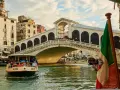 Un vaporetto naviga sul Canal Grande, passando sotto il Ponte di Rialto. L'Italia è rappresentata da una bandiera sventolante.