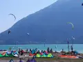 Coloratissima spiaggia sul lago di Santa Croce, con kitesurfisti che sfrecciano sull'acqua turchese e vele colorate che ondeggiano al vento.