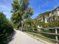 Sentiero ghiaioso lungo il fiume, con alberi verdi e una montagna rocciosa sullo sfondo. La **ciclabile del Brenta** invita a una passeggiata.