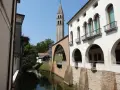 Canale e campanile a Oderzo, cosa vedere: un'elegante facciata bianca con portici e balconi si affaccia sull'acqua, con un ponte ad arco in mattoni.