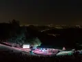 Concerto serale all'anfiteatro del Venda, con un pubblico seduto sull'erba e una band sul palco illuminato, sullo sfondo di un panorama notturno di luci cittadine.