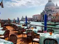 Tavoli apparecchiati con tovaglie azzurre e fiori arancioni, affacciati sul Canal Grande, con gondole e la Basilica di Santa Maria della Salute. Un'ambientazione da sogno per i migliori ristoranti di Venezia.