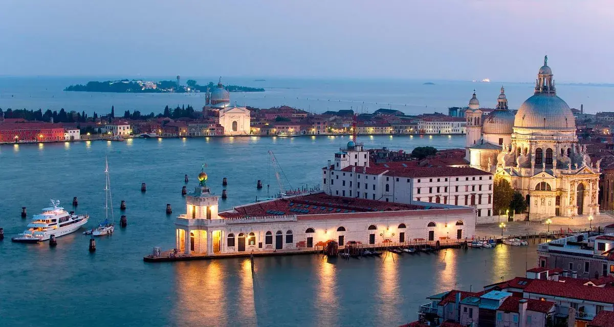 La **Punta della Dogana** a Venezia, con la Basilica di Santa Maria della Salute sullo sfondo, illuminata al crepuscolo. Barche e yacht ormeggiati nell'acqua calma.