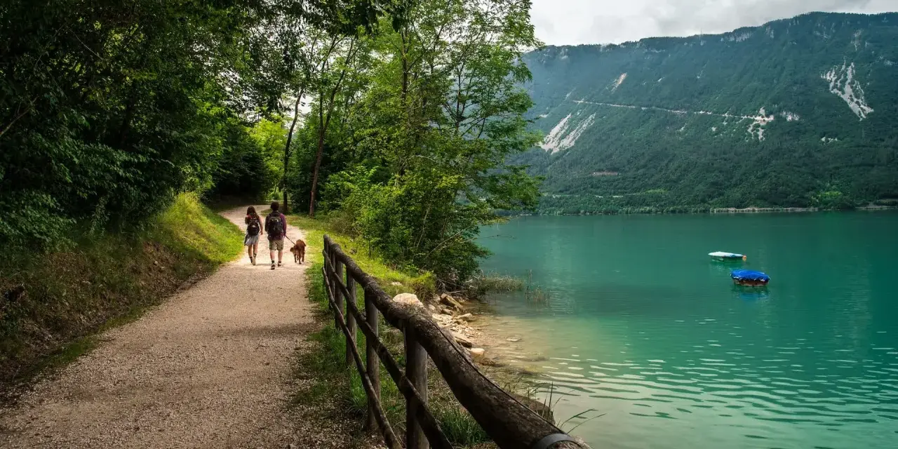 Coppia con cane passeggia lungo il sentiero del lago di Santa Croce, godendosi la natura e il panorama montano.