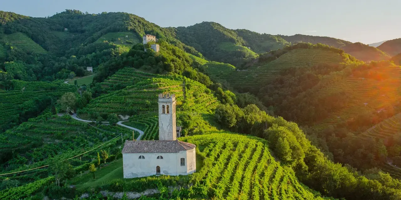Un borgo con una chiesetta e campanile tra colline vitate, un esempio di cosa vedere nei dintorni di Treviso per un paesaggio incantevole.