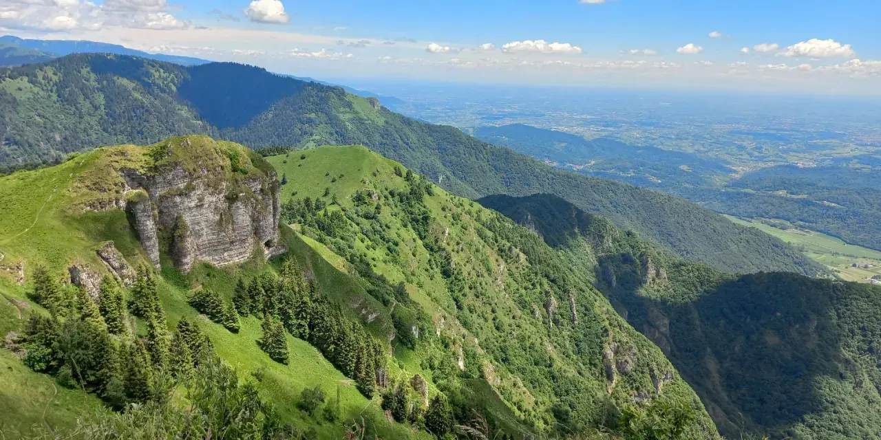 Panoramica mozzafiato dal Passo San Boldo, con verdi pendii montuosi che scendono verso una valle lontana sotto un cielo azzurro con nuvole bianche.