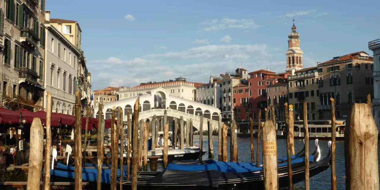 Gondole ormeggiate nel Canal Grande, con il Ponte di Rialto sullo sfondo. Un'immagine di Venezia romantica, con palazzi storici e il cielo azzurro.