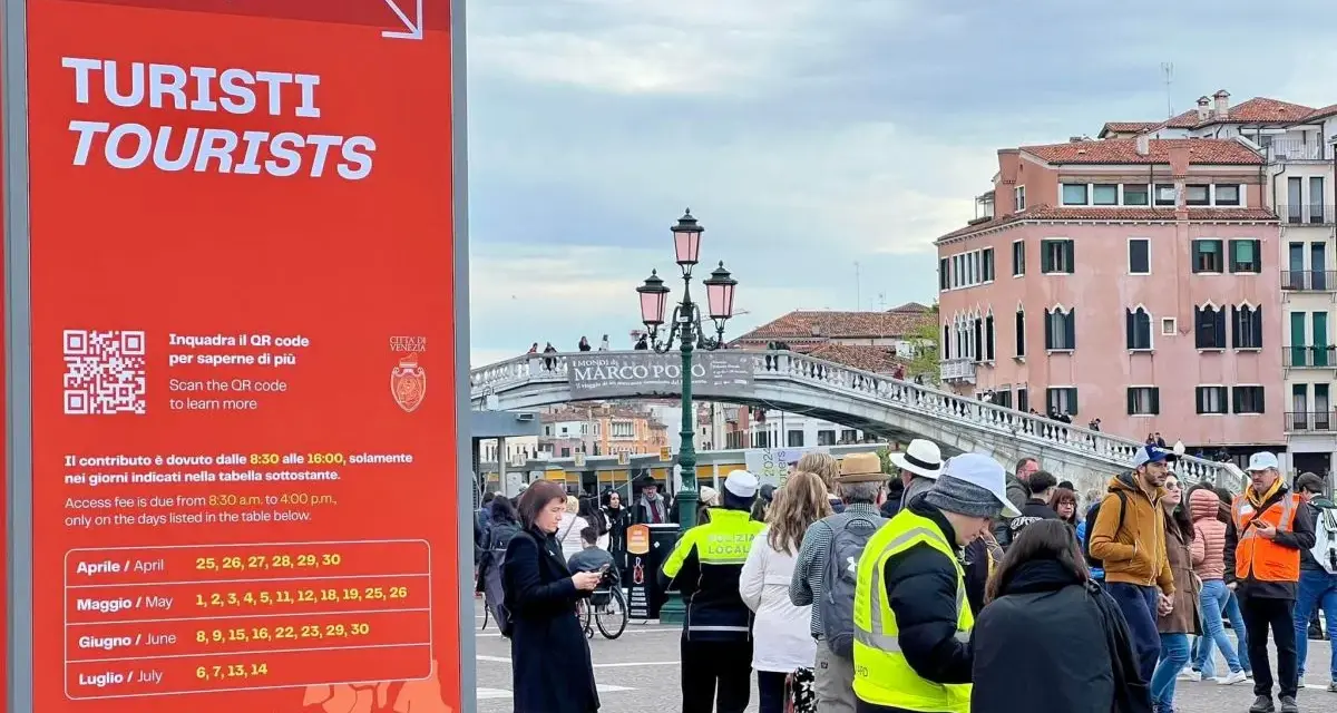 Cartello rosso a Venezia per il contributo accesso turisti, con date e QR code. Persone in fila sullo sfondo.