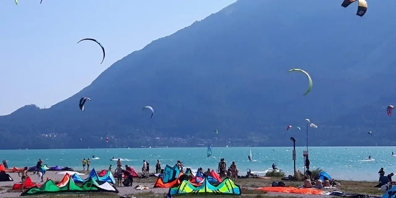 Coloratissima spiaggia sul lago di Santa Croce, con kitesurfisti che sfrecciano sull'acqua turchese e vele colorate che ondeggiano al vento.