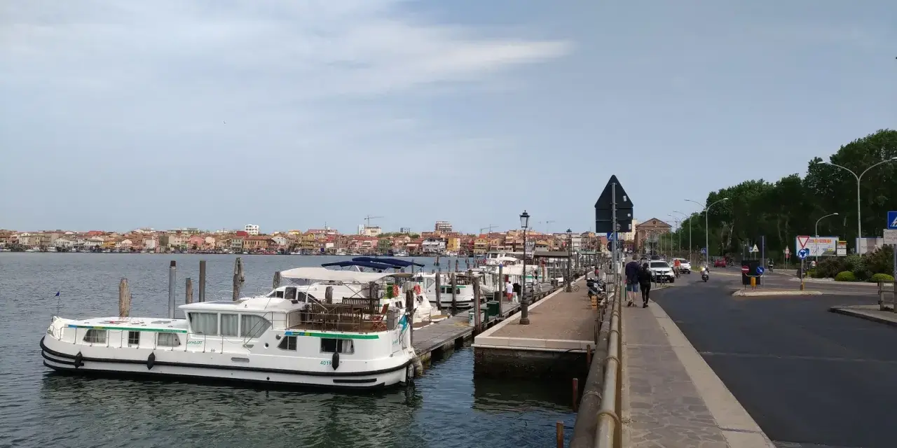 Un vaporetto attracca a Chioggia, con la città sullo sfondo e una strada trafficata.