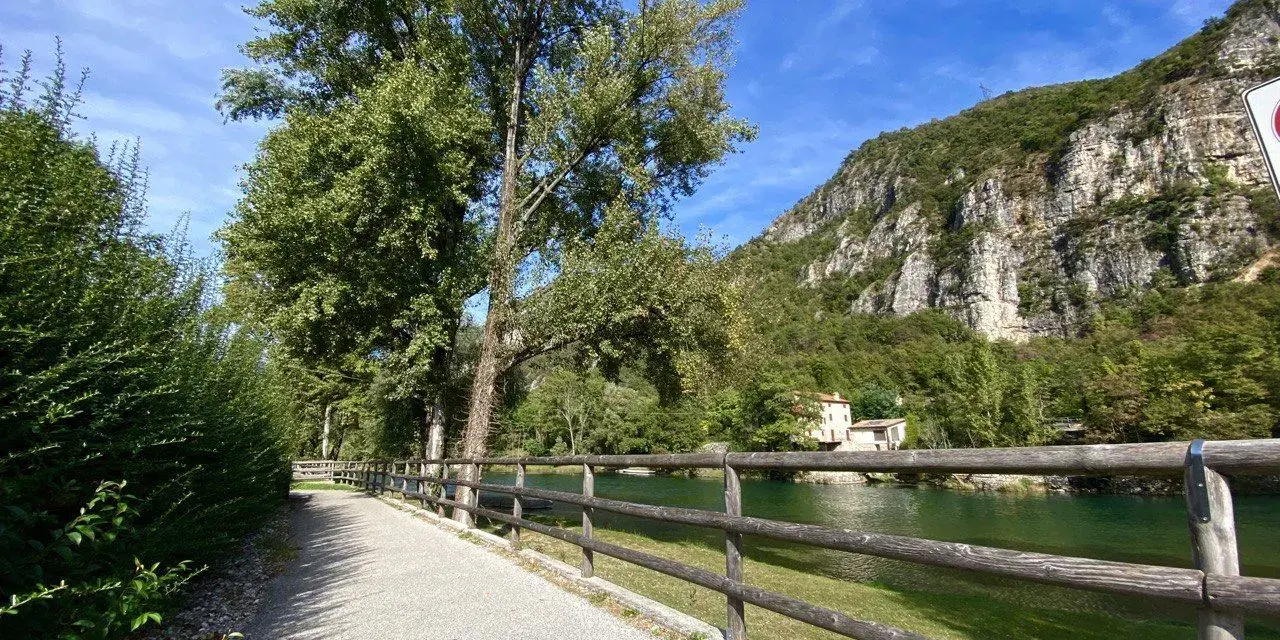 Sentiero ghiaioso lungo il fiume, con alberi verdi e una montagna rocciosa sullo sfondo. La **ciclabile del Brenta** invita a una passeggiata.