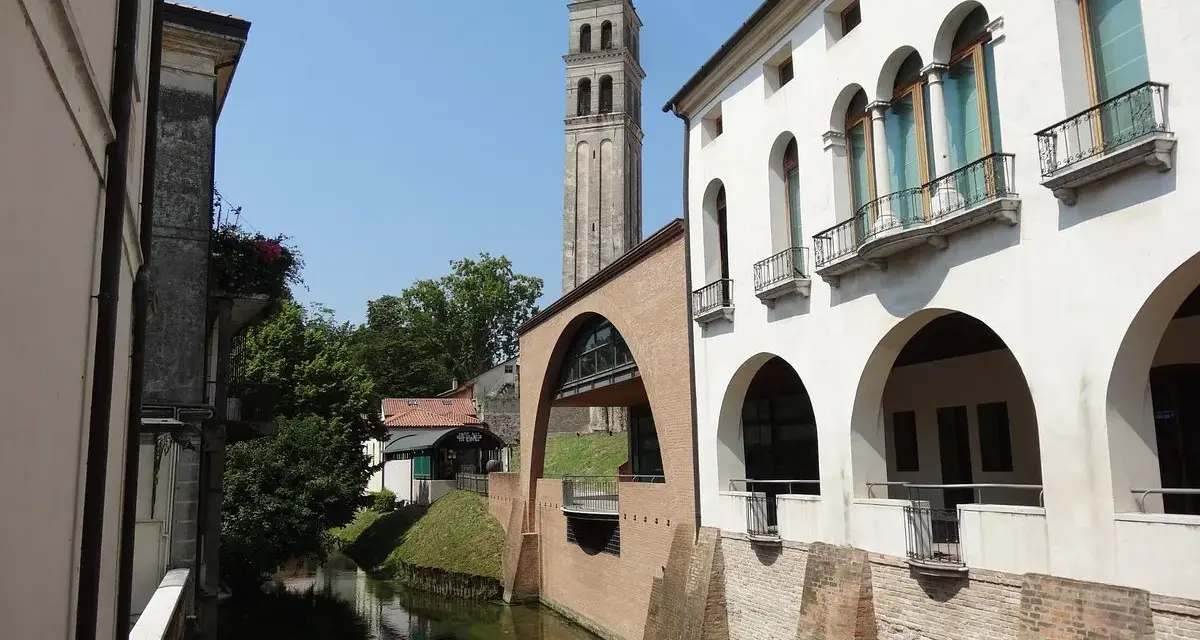 Canale e campanile a Oderzo, cosa vedere: un'elegante facciata bianca con portici e balconi si affaccia sull'acqua, con un ponte ad arco in mattoni.
