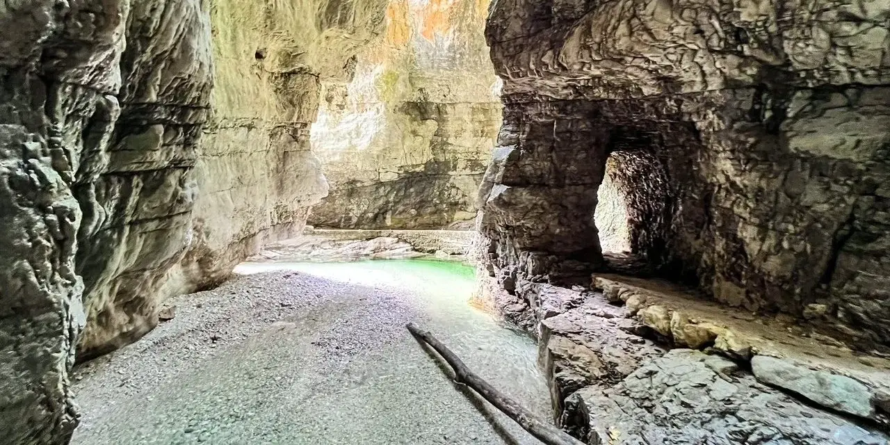 Gola rocciosa con acqua cristallina che ricorda il lago del Mis. Un sentiero scavato nella roccia conduce verso un'apertura luminosa.