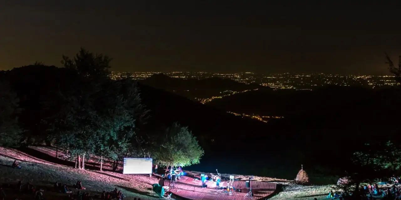 Concerto serale all'anfiteatro del Venda, con un pubblico seduto sull'erba e una band sul palco illuminato, sullo sfondo di un panorama notturno di luci cittadine.
