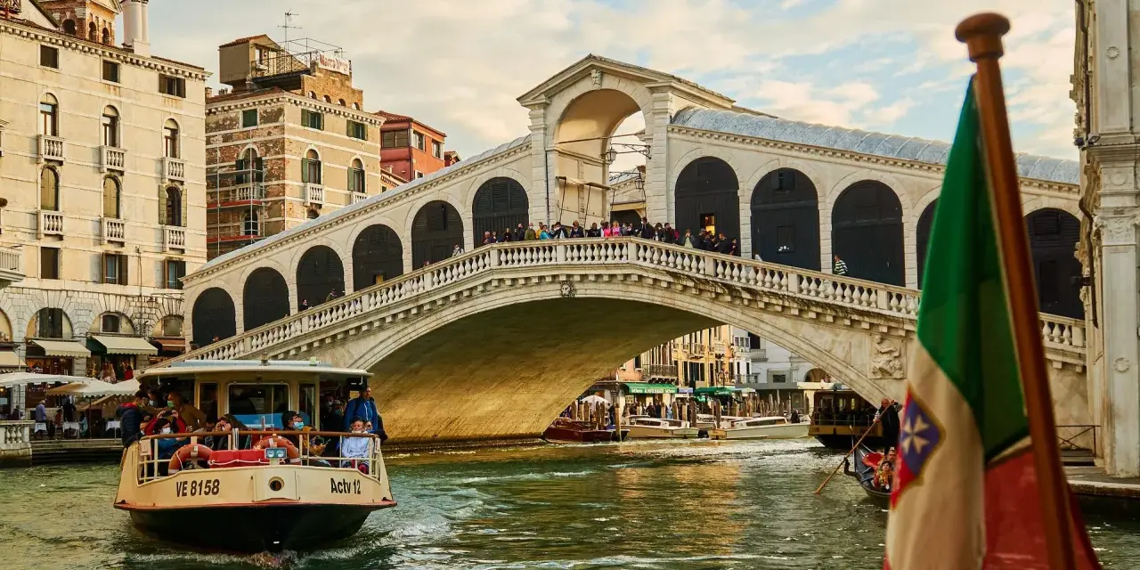 Traghetto della linea 2 di Venezia naviga sul Canal Grande sotto il Ponte di Rialto, con gondole e edifici storici sullo sfondo.