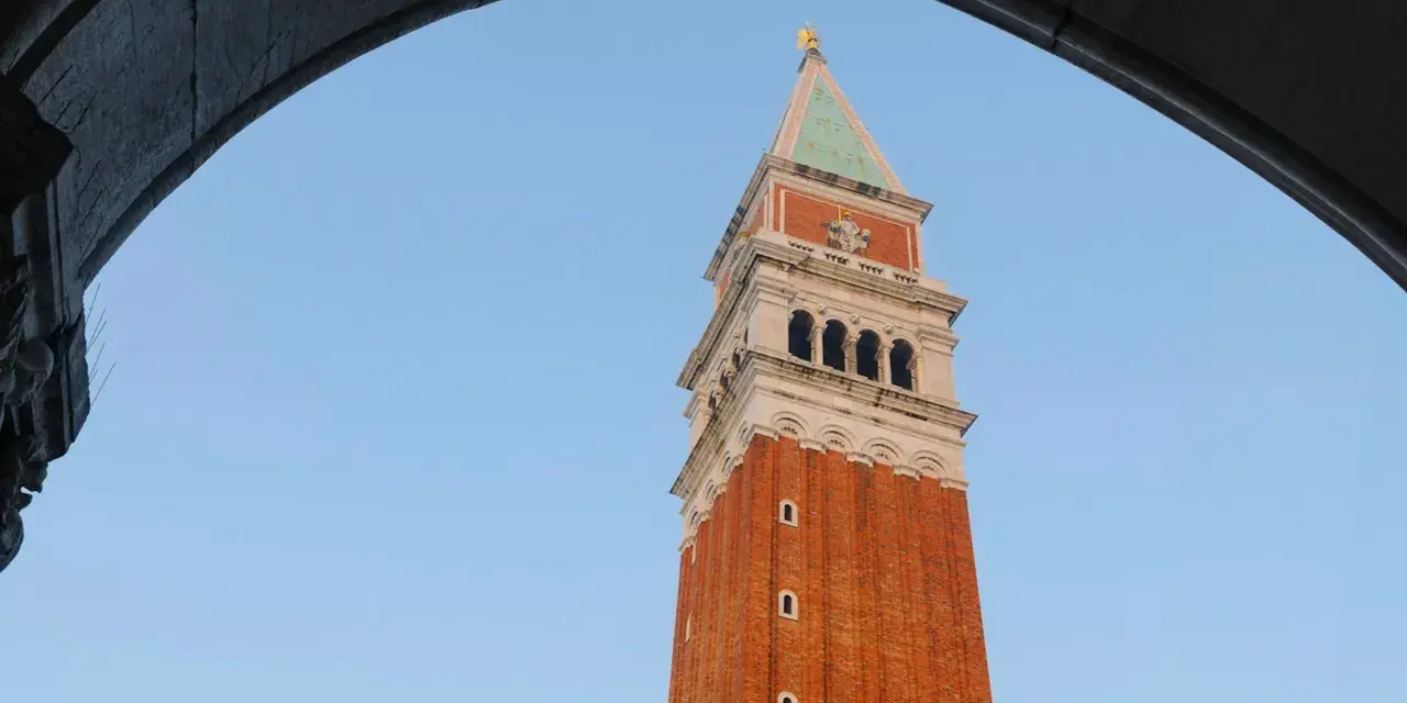 Vista dal campanile di San Marco: la torre in mattoni rossi si staglia contro il cielo azzurro, incorniciata da un arco di pietra.