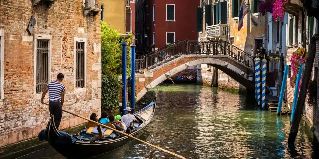 Gondola con turisti naviga in un canale, sotto un ponte, scoprendo la Venezia segreta tra palazzi antichi e fiori.
