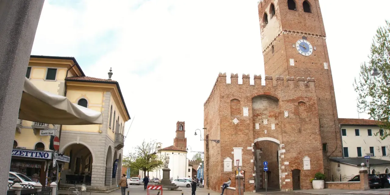 Piazza con torre medievale, portici e negozi. Ecco cosa vedere a Noale: la torre civica con orologio, il palazzo giallo e i tavolini all'aperto.