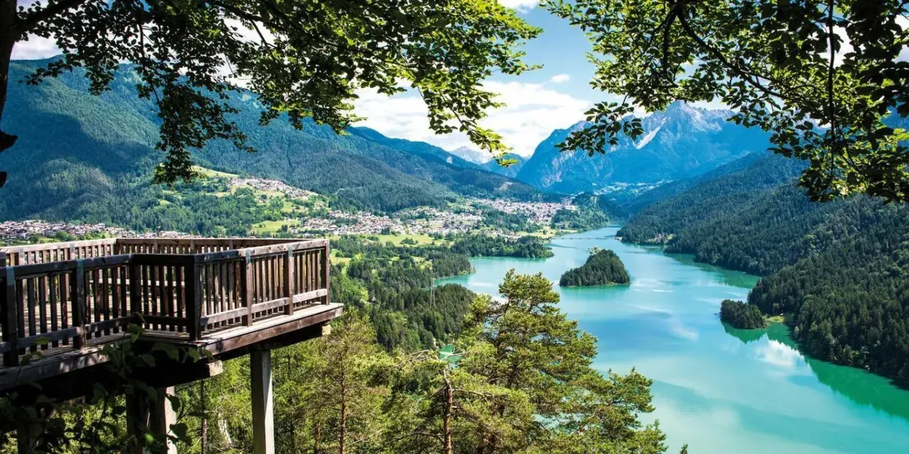 Terrazza panoramica sui laghi del Cadore, con montagne sullo sfondo e un'isola verde nel lago turchese.