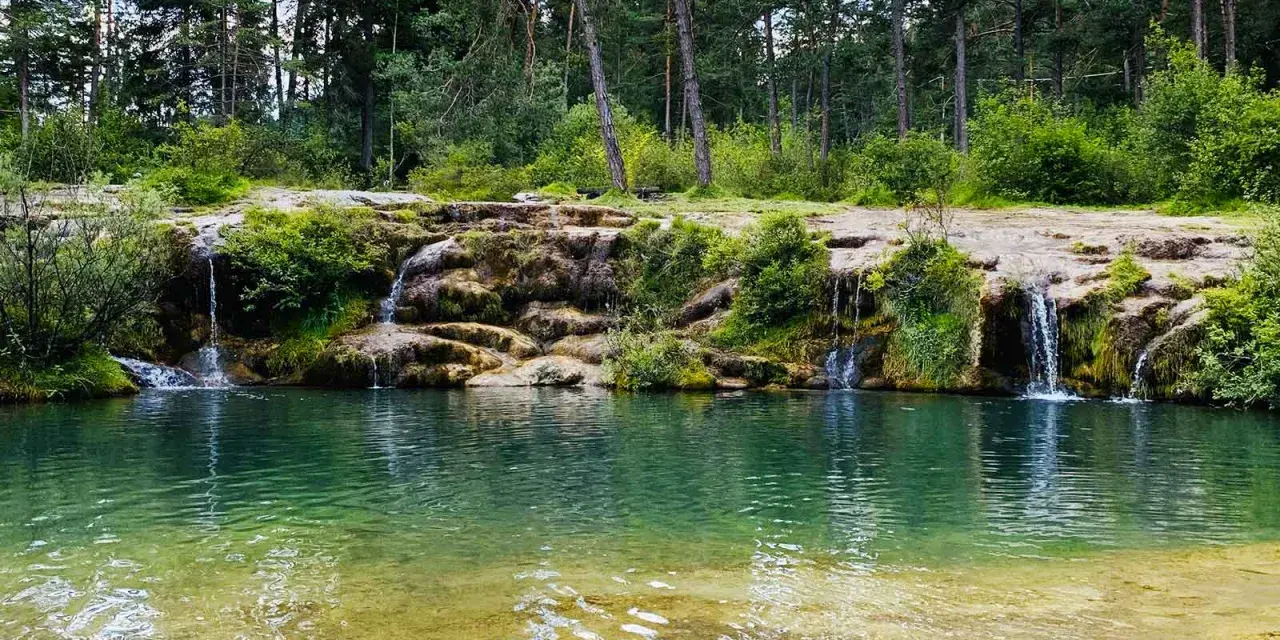 Cascate naturali nella valle di Cadore, con acqua cristallina che scende tra rocce e vegetazione rigogliosa, circondate da un bosco di pini.