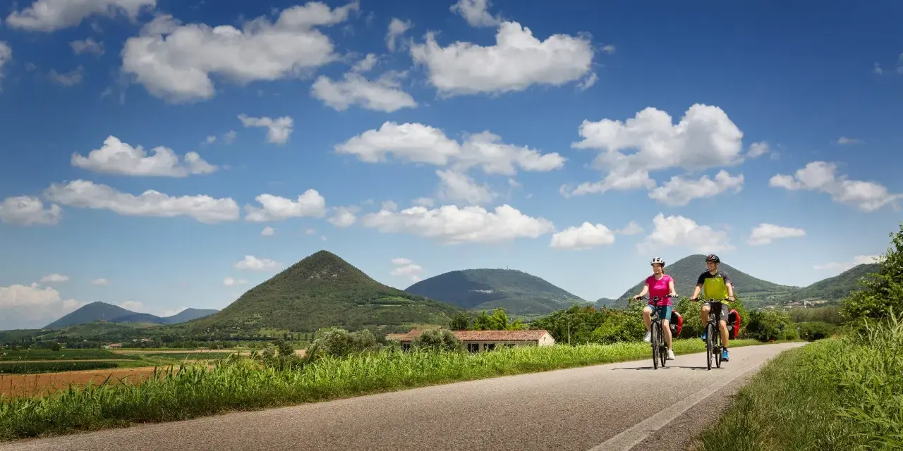 Coppia in bicicletta lungo la ciclabile dei Colli Euganei, tra verdi colline e cielo azzurro con nuvole bianche.