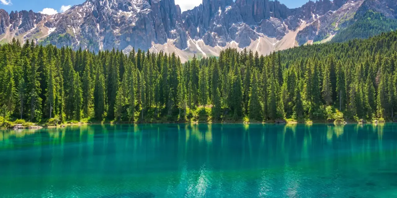 Lago alpino dalle acque turchesi, circondato da una fitta foresta di abeti, con le maestose cime delle Dolomiti innevate sullo sfondo. Un paesaggio da sogno, tra Venezia e le Dolomiti.