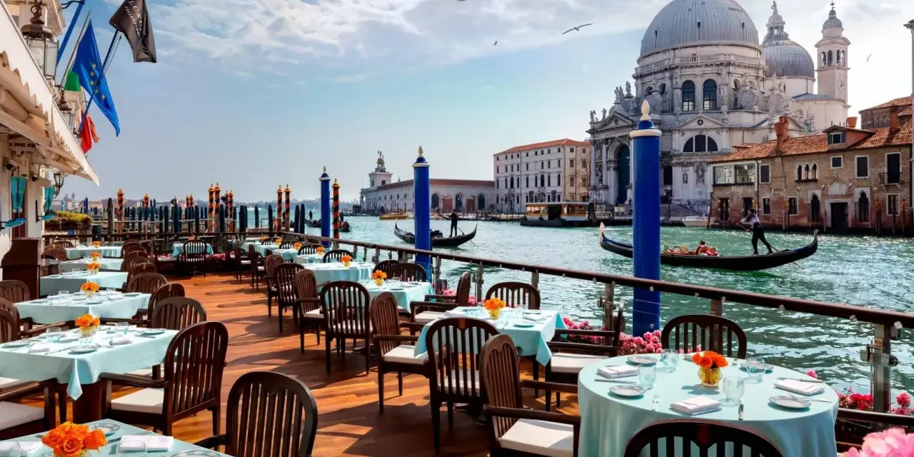 Tavoli apparecchiati con tovaglie azzurre e fiori arancioni, affacciati sul Canal Grande, con gondole e la Basilica di Santa Maria della Salute. Un'ambientazione da sogno per i migliori ristoranti di Venezia.