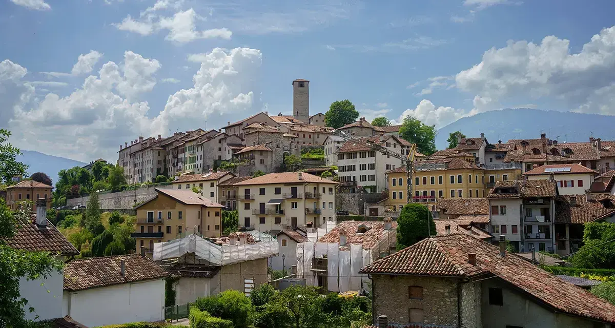 Panoramica di Feltre e dintorni, con case dai tetti rossi che si arrampicano sulla collina sotto un cielo azzurro e nuvoloso.