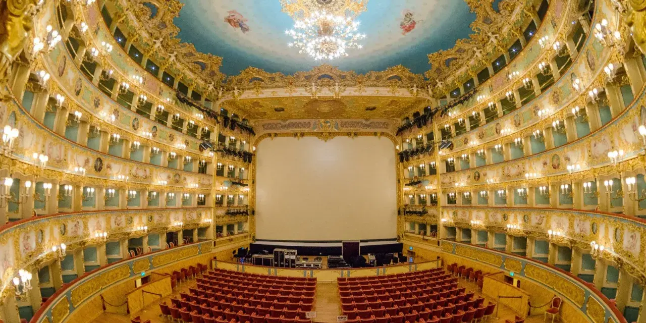 Interno sfarzoso del Teatro La Fenice di Venezia, con palchi dorati, soffitto affrescato e un grande schermo bianco.