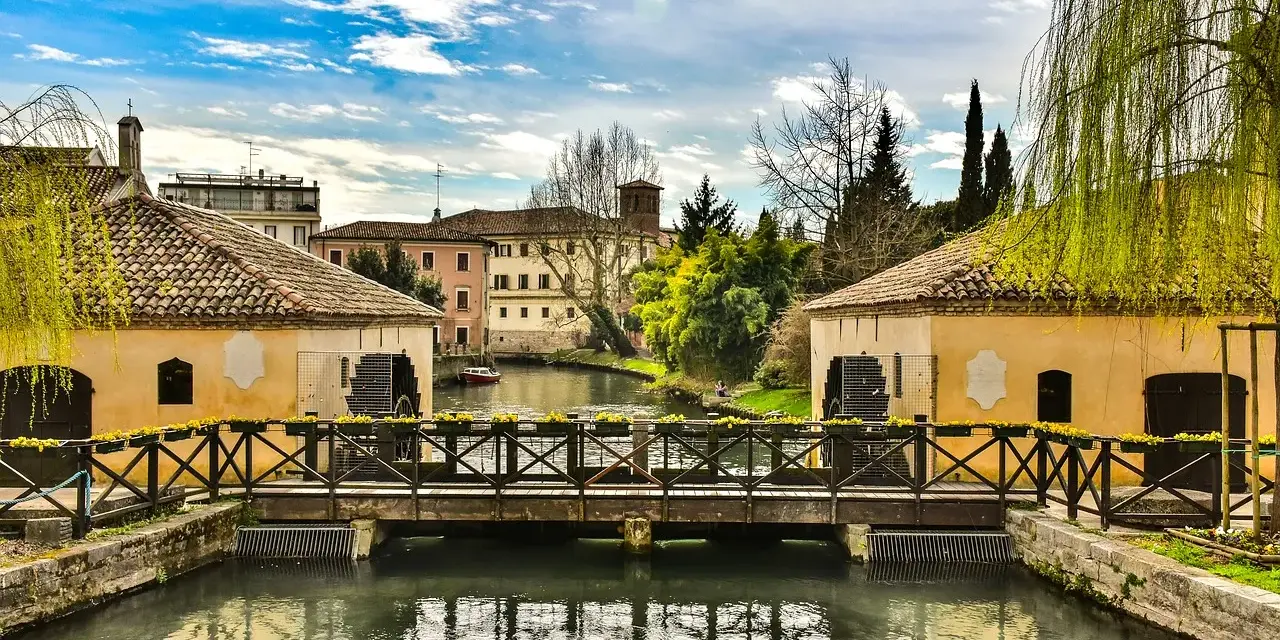 Ponte in legno su un canale con mulini ad acqua, case storiche e vegetazione rigogliosa, un'atmosfera serena che ricorda Portogruaro Caorle.