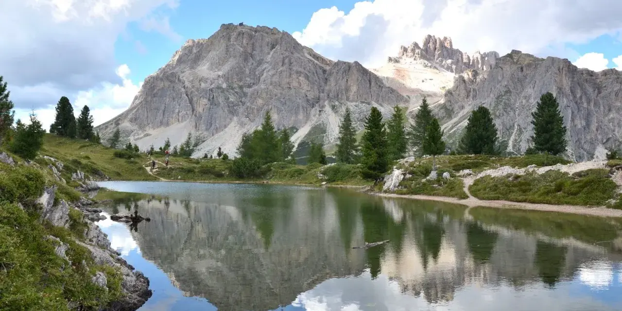 Riflessi perfetti delle montagne nel **lago di Limedes**, con sentiero e pini che incorniciano il paesaggio.