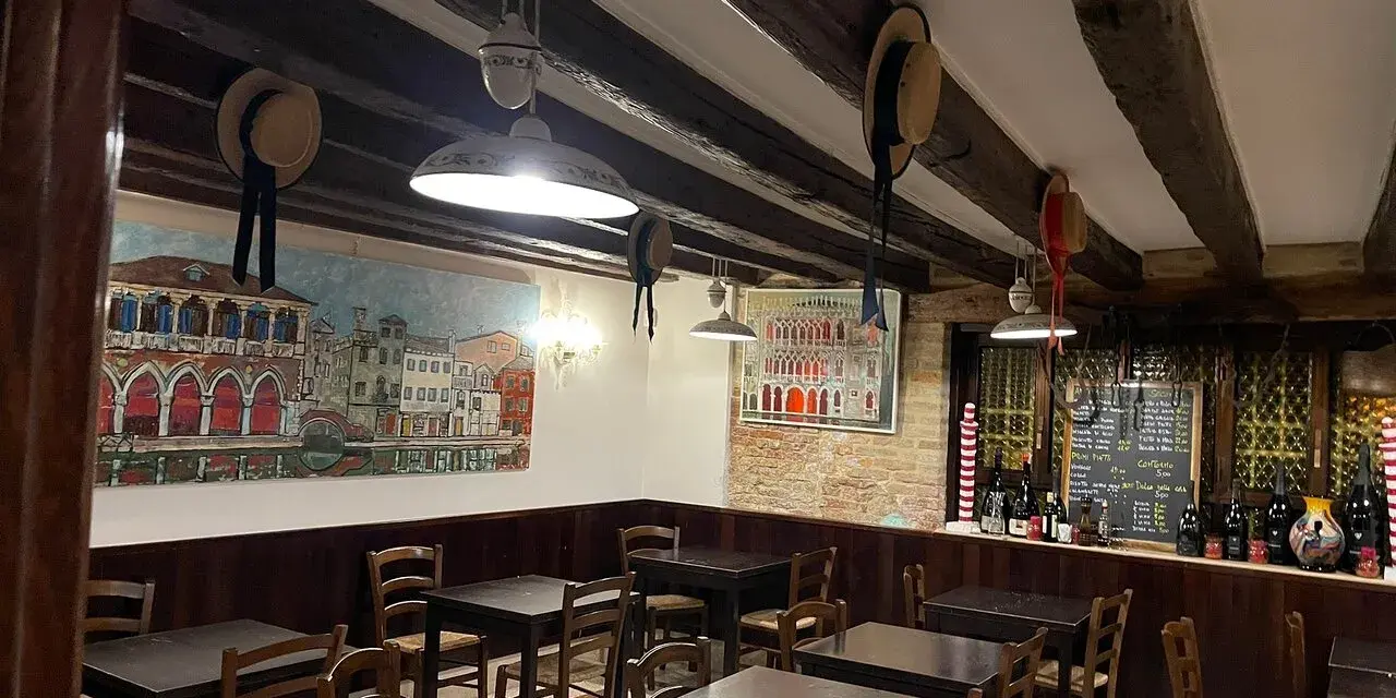 Interno di un **ristorante antico** con dipinti di **Venezia**, tavoli in legno e un calice di vino.