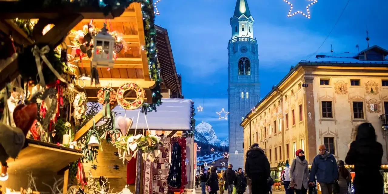 Mercatino di Natale a Cortina d'Ampezzo, con bancarelle addobbate, luci scintillanti e la torre del campanile che domina il cielo serale.