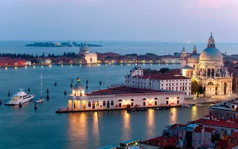 La **Punta della Dogana** a Venezia, con la Basilica di Santa Maria della Salute sullo sfondo, illuminata al crepuscolo. Barche e yacht ormeggiati nell'acqua calma.