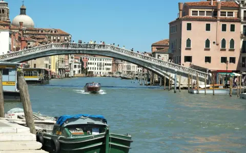 Un motoscafo naviga sotto uno dei ponti di Venezia, con edifici storici e gente che passeggia.