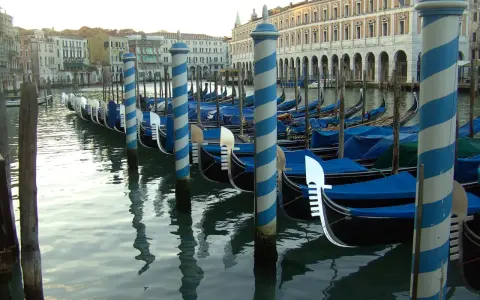 Una fila di imbarcazioni tipiche veneziane, le gondole, ormeggiate lungo un canale, con i loro caratteristici pali a strisce blu e bianche.