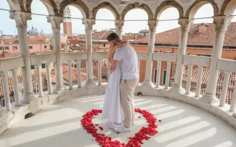 Un uomo abbraccia una donna su un balcone con vista su Venezia, circondati da petali di rosa a forma di cuore. Una perfetta proposta matrimonio Venezia.