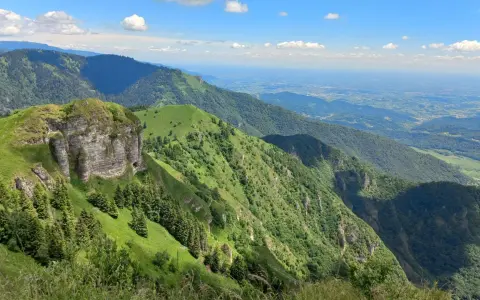 Panoramica mozzafiato dal Passo San Boldo, con verdi pendii montuosi che scendono verso una valle lontana sotto un cielo azzurro con nuvole bianche.