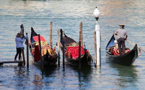 Gondole veneziane ornate, una turista scatta foto. La differenza tra gondola e sandalo è chiara: una naviga, l'altro si indossa.