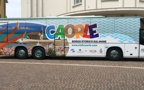 Bus turistico con grafica promozionale di Caorle, borgo storico sul mare. Immagini di spiaggia, mare e una donna in bikini.