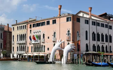 Enorme installazione di mani bianche emerge dal Canal Grande, davanti al museo arte contemporanea Venezia. Gondole ormeggiate.
