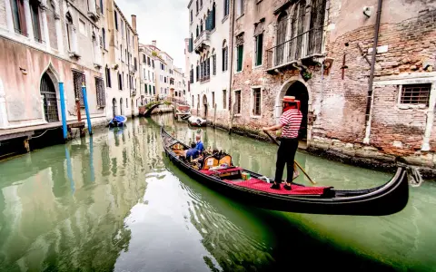 Gondoliere in abito a righe guida una gondola con turisti lungo un canale veneziano. Chissà quanto costa un giro in gondola a Venezia per vivere questa magia.