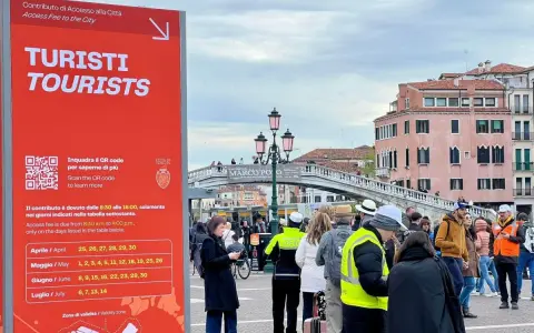 Cartello rosso a Venezia per il contributo accesso turisti, con date e QR code. Persone in fila sullo sfondo.