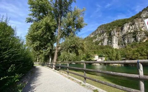 Sentiero ghiaioso lungo il fiume, con alberi verdi e una montagna rocciosa sullo sfondo. La **ciclabile del Brenta** invita a una passeggiata.