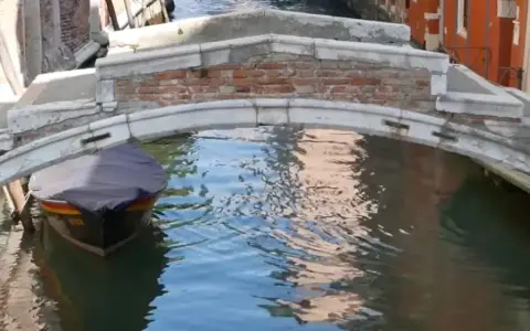 Un piccolo ponte a Venezia, senza parapetto, sovrasta un canale dove una barca coperta attende.