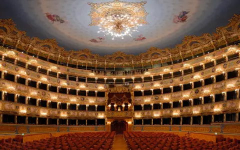 Interno sfarzoso del teatro La Fenice a Venezia, con palchi dorati, affreschi sul soffitto e un lampadario scintillante.