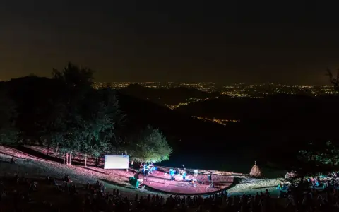 Concerto serale all'anfiteatro del Venda, con un pubblico seduto sull'erba e una band sul palco illuminato, sullo sfondo di un panorama notturno di luci cittadine.