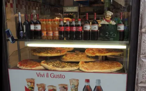 Vetrine con pizze, bottiglie di vino e bibite, un cartello "PIZZA DA € 2.50" e un cuoco decorativo. Sembra un ottimo posto per trovare le migliori pizzerie a Venezia.