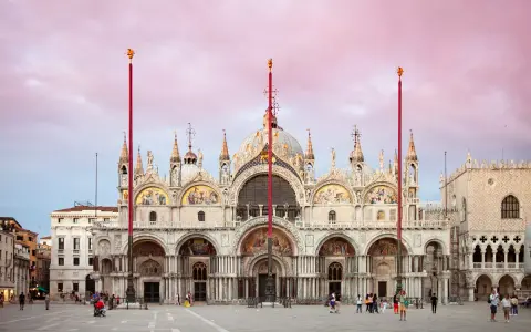 La maestosa basilica di San Marco a Venezia, con le sue cupole e mosaici dorati, si staglia sotto un cielo rosa al tramonto.